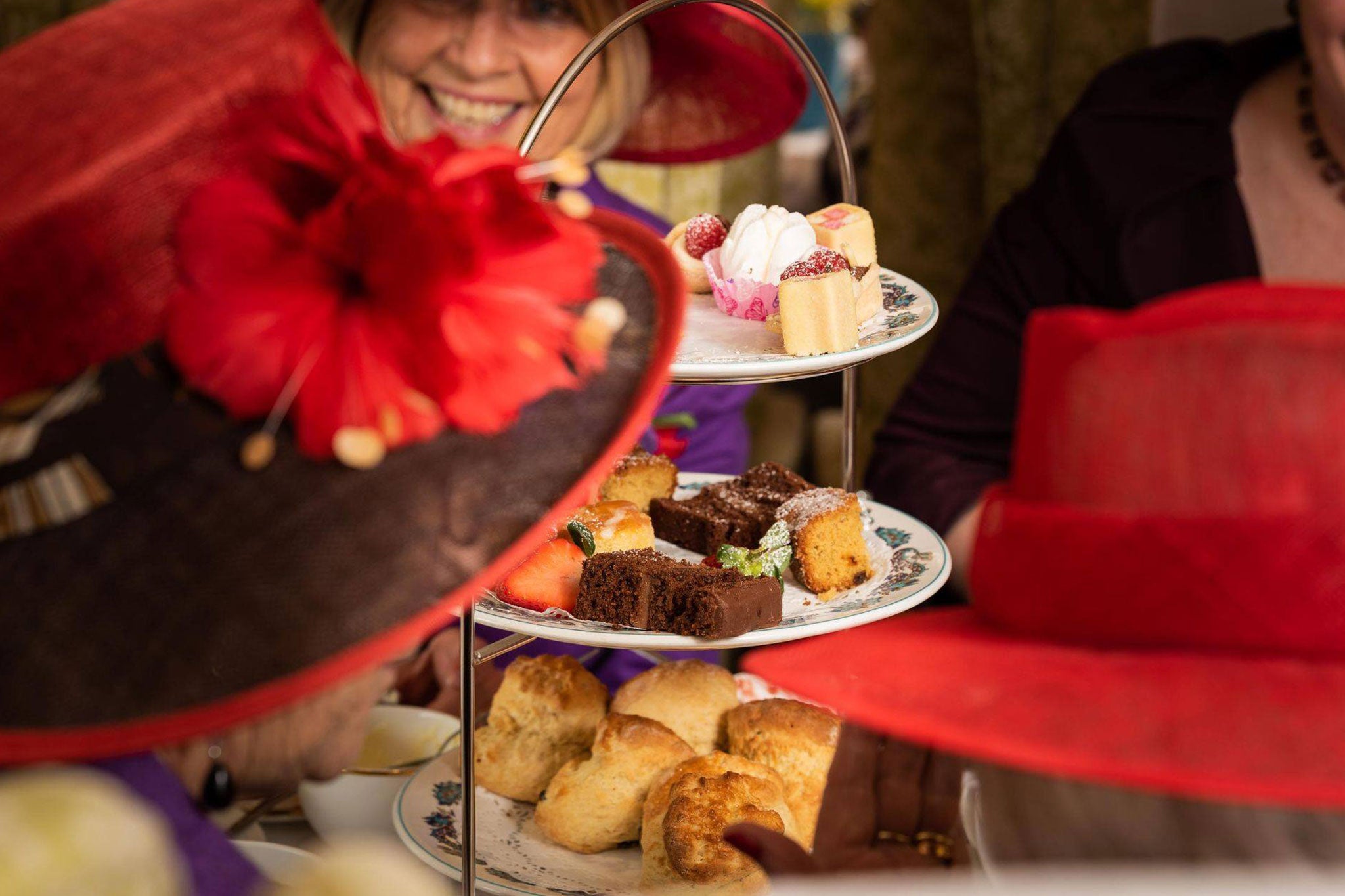 People wearing red hats with floral decorations at a high tea event, with pastries on a tiered stand.